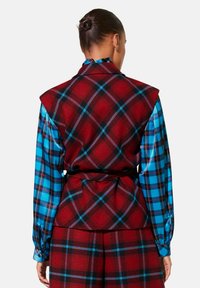 Rood en zwart geruite vest met scherpe schouders, gecombineerd met een blauw en zwart geblokt overhemd. Het materiaal lijkt dik en gestructureerd.