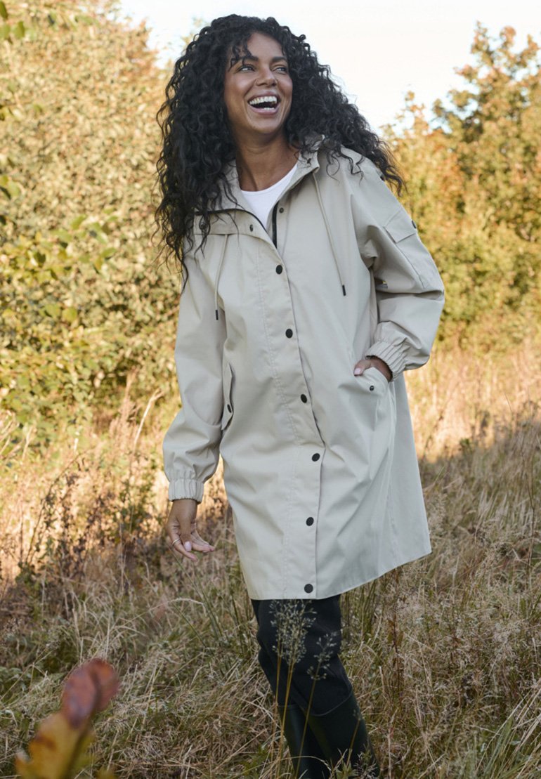 Beige lightweight jacket with a drawstring hood, snap buttons, and elastic cuffs, worn over a white top, in a grassy outdoor setting.