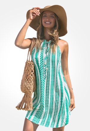 Mujer sonriente, vestida con un vestido sin mangas de patrón verde y blanco, sombrero de ala ancha y llevando un bolso tejido al hombro.