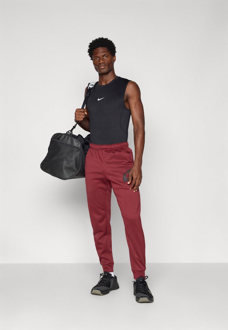 Top senza maniche nero, pantaloni tuta bordeaux con logo e scarpe da ginnastica nere. Tiene una borsa da palestra nera, in piedi su uno sfondo semplice.