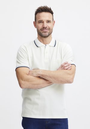 Hvit polo-skjorte med marineblå kant på kragen og ermene, med en liten brystlomme og en rød og hvit logo aksent.