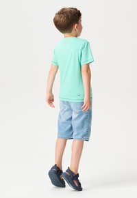 Miętowa koszulka z krótkim rękawem, niebieskie szorty dżinsowe i granatowe sandały. Metka z tyłu koszulki. Dziecko widziane z tyłu.