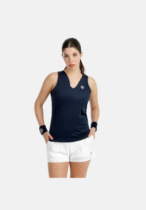 Junge Frau in ärmellosem marineblauem Sporttop und weißen Shorts, trägt schwarze Handgelenkbandagen, steht mit den Händen in den Taschen vor einfachem Hintergrund.