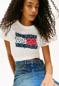 T-shirt blanc en coton avec un graphique floral et un drapeau en bleu, rouge et blanc. Associé à une jupe en denim bleue taille haute.