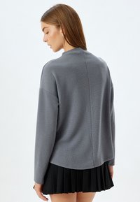 Maglione grigio strutturato con un collo alto e spalle scese, abbinato a una gonna nera plissettata, visto da dietro.
