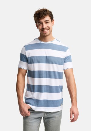 Ung mand smilende, iført en hvid og blåstribet kortærmet skjorte og lysegrå jeans, stående foran en ensfarvet baggrund.