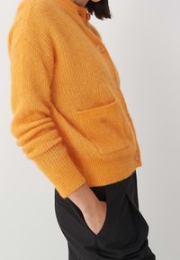 Orange ullkofta med rund hals, långa ärmar och två framfickor. Har tre knappar och en mjuk, texturerad yta.