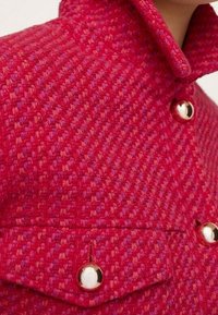 Czerwona tkana kurtka z wysokim kołnierzem, złotymi guzikami i dwiema kieszeniami na piersi z fakturowanym wzorem.
