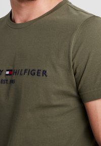 Mand iført olivengrøn Tommy Hilfiger t-shirt med broderet logo og tekst på brystet, hvor kortærmerne er synlige.
