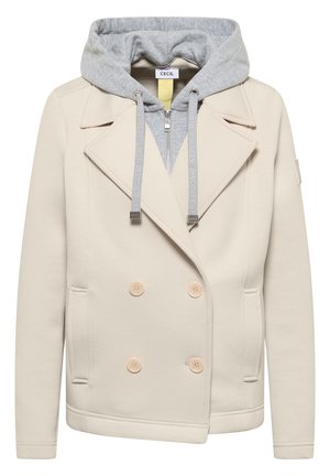 Beżowa dwurzędowa marynarka z czterema guzikami i przednimi kieszeniami, nałożona na jasnoszarą bluzę z kapturem zapinaną na zamek, z troczkami.