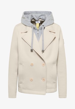 Beżowa dwurzędowa marynarka z czterema guzikami i przednimi kieszeniami, nałożona na jasnoszarą bluzę z kapturem zapinaną na zamek, z troczkami.
