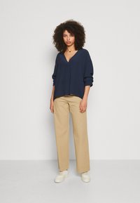 Blusa blu navy a vestibilità rilassata con scollo a V e maniche arrotolate, abbinata a pantaloni a gamba larga beige chiaro e sneaker bianche.