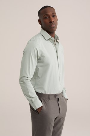 Muž nosi svetlozelenú košeľu na gombíky a sivé nohavice, stojí s rukami v vreckách pred jednofarebným pozadím.