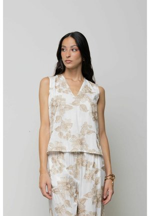 Donna con lunghi capelli scuri che indossa un top bianco senza maniche e pantaloni con motivo floreale beige, in piedi davanti a uno sfondo chiaro uniforme.