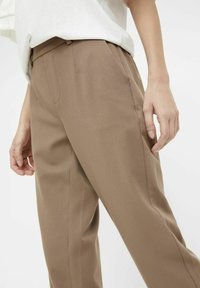 Bruine op maat gemaakte broeken van textuurstof, met zijzakken en een rechte pijp. G gedragen met een witte losse top.