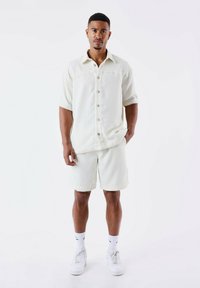 Camisa de manga corta con cuello y pantalones cortos, ambos en tejido de color crema texturizado. Presenta grandes botones y un ajuste relajado, combinados con zapatillas blancas.