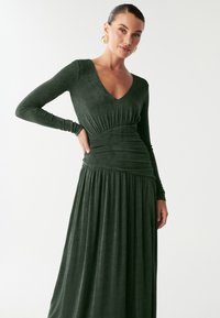 Dlhé zelené šaty s výstrihom do V, s detailom nariasenej pása a prúdnou sukňou. Mäkká tkanina s jemnou textúrou. Zlaté náušnice dotvárajú vzhľad.