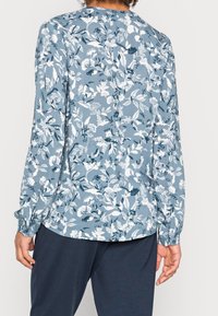 Blusa floreale di un blu chiaro con modelli di fiori bianchi e blu scuro. Maniche lunghe con polsini arricciati, orlo arrotondato e vestibilità comoda.