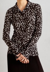 Chemise noire à manches longues avec un motif abstrait blanc, col pointu, associée à une jupe plissée noire.