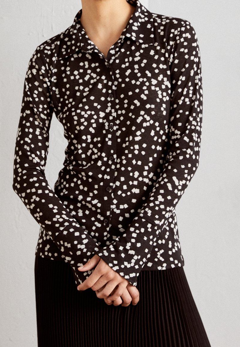Chemise noire à manches longues avec un motif abstrait blanc, col pointu, associée à une jupe plissée noire.