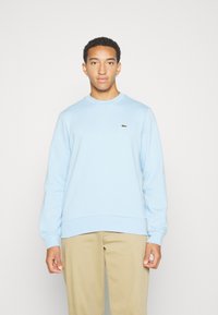 Sweatshirt bleu clair avec un col et des poignets côtelés, affichant un petit logo vert sur la poitrine, fabriqué dans un tissu doux, coupe décontractée.