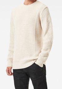 Light beige knit sweater with a ribbed texture, featuring patterned sleeves and a classic crew neckline, paired with black trousers.