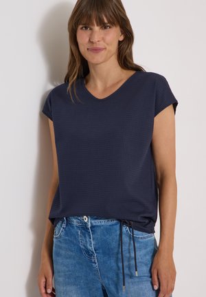 Femme aux cheveux bruns et lisses portant un haut bleu marine à manches courtes et un jean bleu, debout contre un mur clair uni.