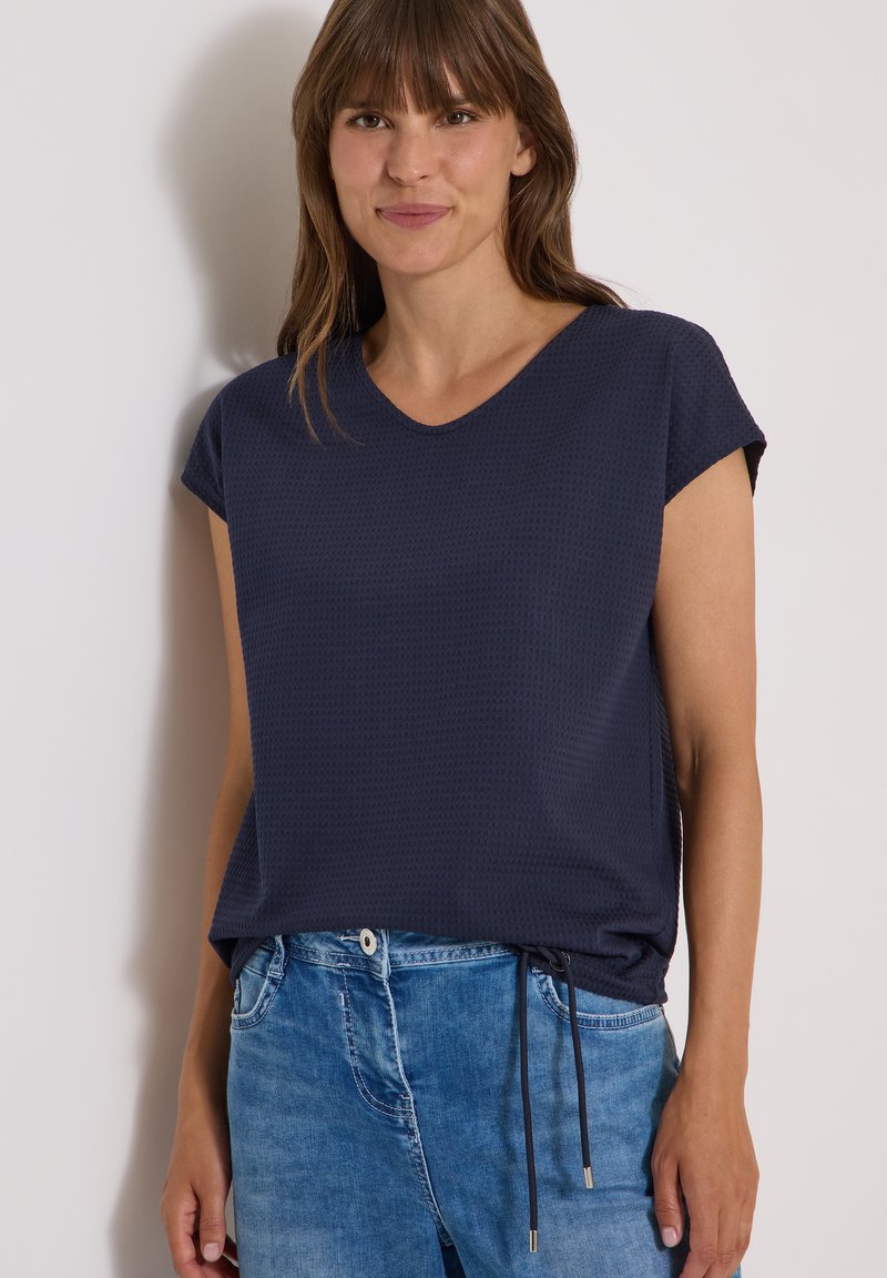 Femme aux cheveux bruns et lisses portant un haut bleu marine à manches courtes et un jean bleu, debout contre un mur clair uni.