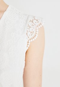 Hvit bluse i blonder med bølget erme som har intrikate blomster mønstre og perle dekorer langs kanten. Myk tekstur er tydelig.