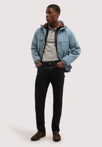 Hombre joven con las manos en los bolsillos, vistiendo una chaqueta con capucha azul claro, camisa polo gris, pantalones negros y mocasines marrones, mirando hacia un lado.