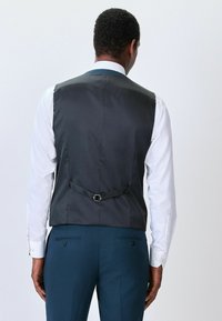 Dunkelgraue Weste mit glatter Textur, ausgestattet mit einem einzelnen Riemen und einer Schnalle auf der Rückseite, kombiniert mit einem langärmeligen weißen Hemd.