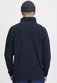 Sudadera de fleece color marino con cuello alto, textura suave y mangas largas. Se lleva con un gorro de punto oscuro en la parte superior.