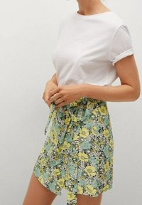 Jupe portefeuille florale verte ornée de fleurs jaunes et blanches, associée à un t-shirt blanc uni. La jupe présente une taille nouée.