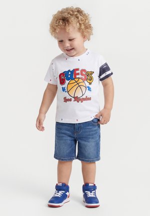 Peuter met krullend haar die een wit basketbalthema-shirt, spijkershorts en blauwe hoge sneakers draagt, glimlacht en kijkt naar beneden.