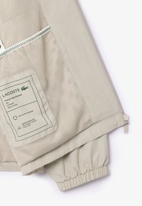 Lacoste hellbeige Sportjacke mit Netzfutter, elastischen Manschetten, einem Etikett aus recyceltem Polyester und wasserabweisenden Details.