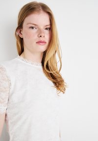 Hvit blonde topp med bølget halskant og korte gjennomsiktige ermer, med et teksturert blomsterdesign og et jevnt, lett stoff.