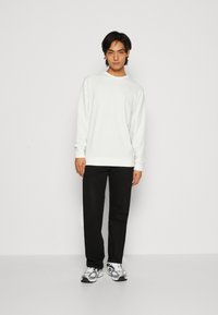 Hvid langærmet sweatshirt med ribbede manchetter og kant, parret med sorte bukser og sølvfarvede sneakers, stående mod en ensfarvet baggrund.
