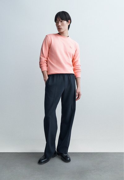 Νεαρός άνδρας στέκεται με τα χέρια στις τσέπες, φοράει ροδακινί φούτερ, μαύρο παντελόνι με φαρδύ πόδι και μαύρα παπούτσια, σε απλό ανοιχτό φόντο.