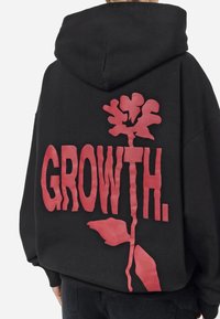 Sweat à capuche noir avec un large motif rouge d'une fleur stylisée et le mot « GROWTH. » imprimé verticalement dans le dos.