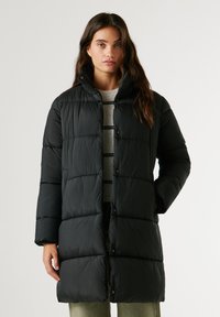 Melnā pūkainā jaka ar kapuci, ar kvadrātveida šuvēm, spiedpogām un sānu kabatām. Audums izskatās gluds un viegls.