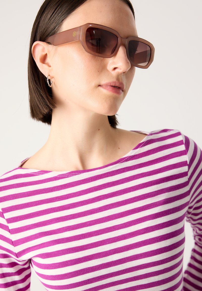 Braune quadratische Sonnenbrille mit einem durchsichtigen Gestell, getragen mit einem lila-weiß gestreiften Langarmshirt. Dezenter Ohrstecker sichtbar.