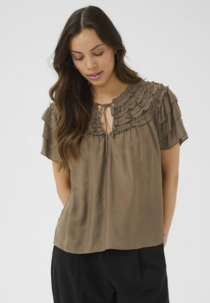 Brown, short-sleeved blouse with ruffled neckline and tie detail. Smooth texture with a relaxed fit. Worn with black trousers.