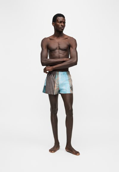 Homme torse nu debout pieds nus, les bras croisés, portant un short de bain rayé bleu clair et marron, regardant sur le côté sur fond blanc.