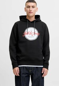 Black hoodie made from smooth fabric featuring a circular graphic with text. Drawstrings are black and white. Paired with dark denim.