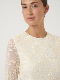 Νέα γυναίκα φορώντας μια διάφανη λευκή μπλούζα με κεντημένα λουλουδάτα σχέδια και μικρές σκουλαρίκια κρίκους σε απλό φόντο.