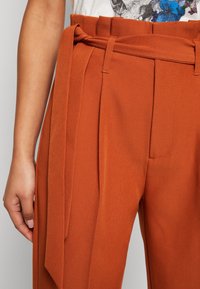 Rostfärgade byxor med hög midja, som har ett brett bälte, en plisserad design och en mjuk textur. Detaljerad sömnad på framsidan.