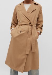 Beige kaksirivinen trenssitakki, jossa on vyötärönauha, leveät kaulukset ja käännetyt hihat. Kangas näyttää sileältä, ja takissa on etutaskut.