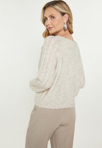 Maglione lavorato a maglia beige chiaro con una vestibilità ampia, caratterizzato da motivi testurizzati sulle maniche. Materiale morbido e accogliente con un collo casual.