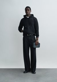 Mladý muž v čiernej mikine s kapucňou a širokých nohaviciach, držiaci malú čiernu kabelku, stojaci pred jednoduchým sivým pozadím.