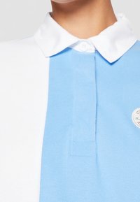 Blå och vit polo skjorta med krage, har en knappknäppning och en broderad patch på bröstet. Mjuk bomullstextur.